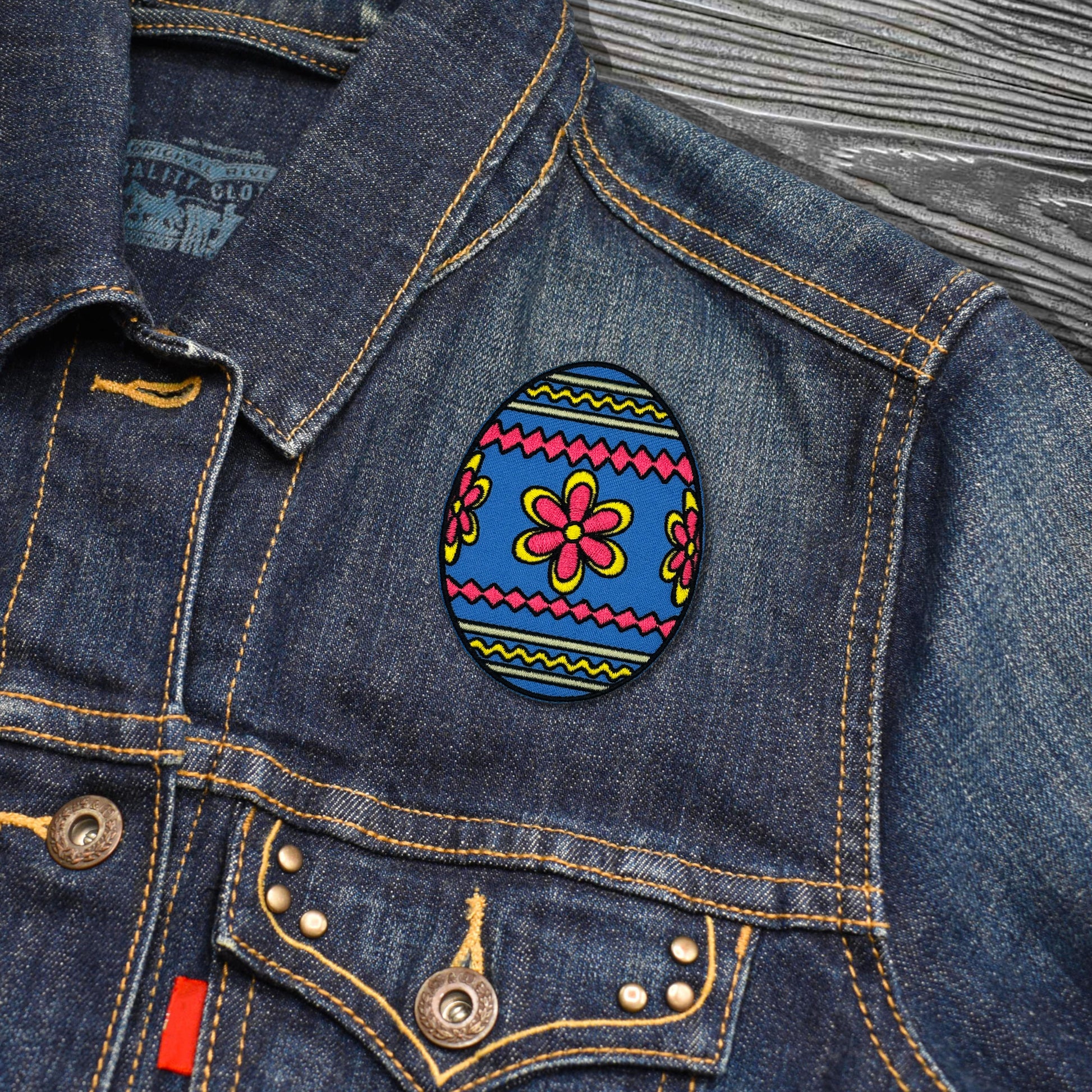 Denim jacket with a colorful floral Easter Egg patch on a wooden surface
