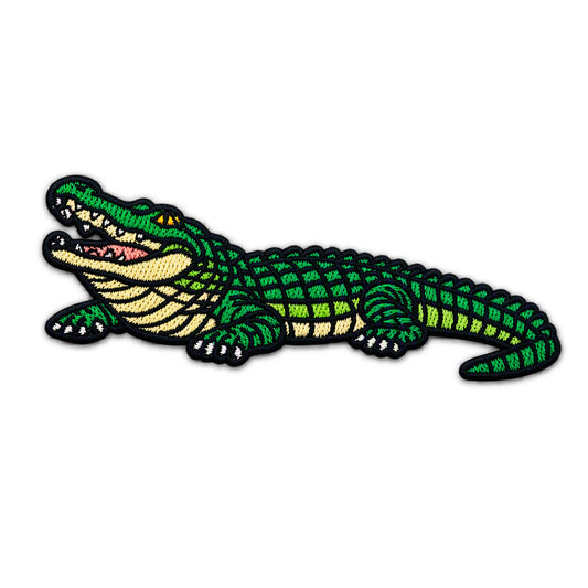 An embroidered iron-on patch of a cartoon alligator with an open mouth, featuring green scales, a beige underbelly, and black stitched outlines on a white background.