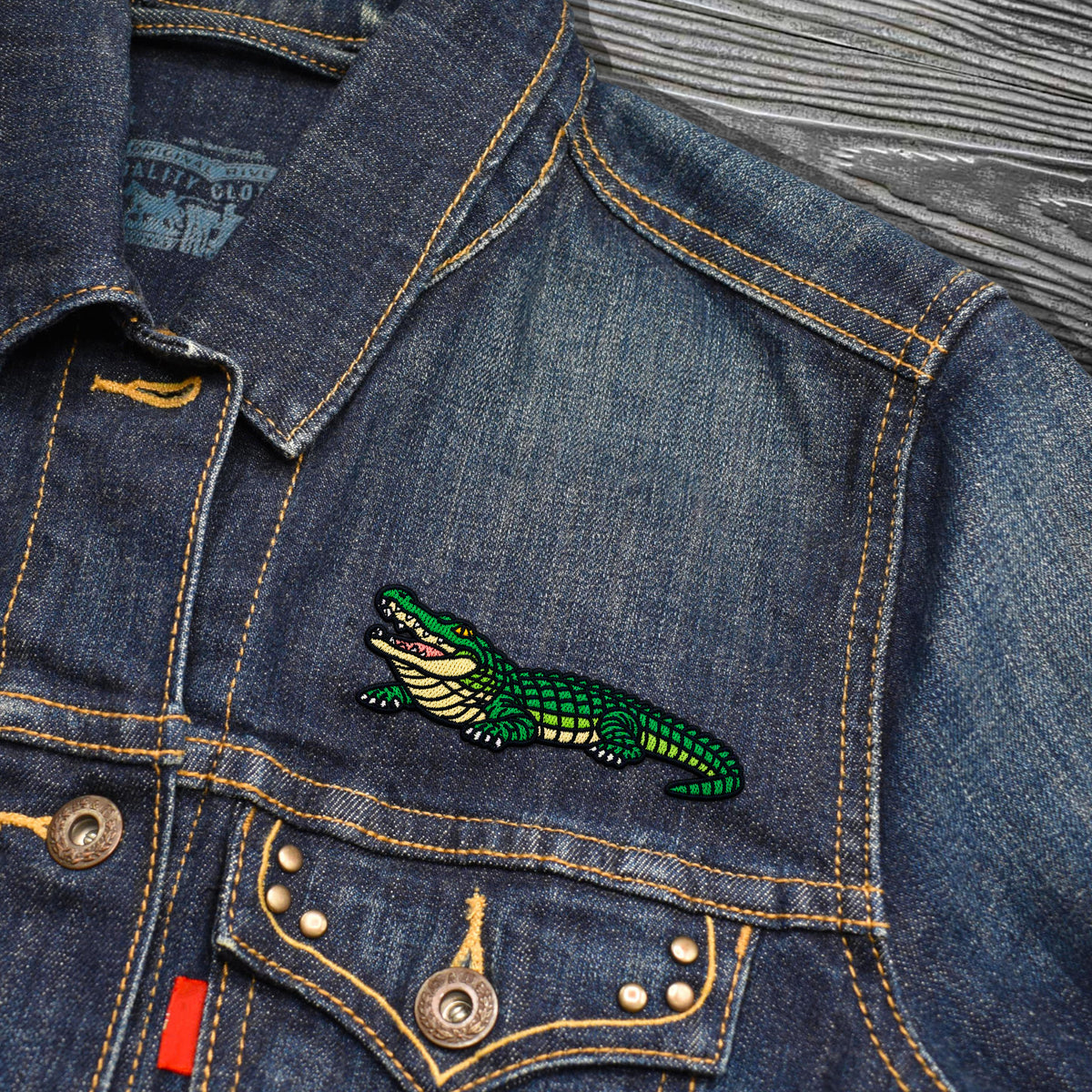 Close-up of a blue denim jacket with a green embroidered alligator patch on the chest pocket, showing yellow contrast stitching and metal buttons.
