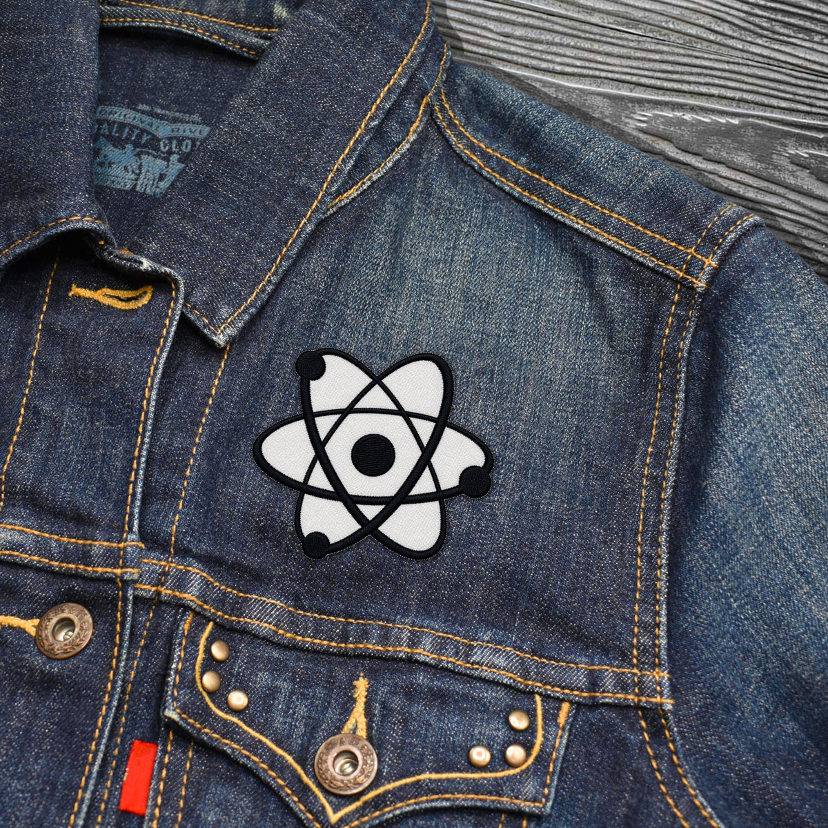 A close-up of a blue denim jacket featuring a white and black atom patch on the chest, with yellow contrast stitching and metal buttons.