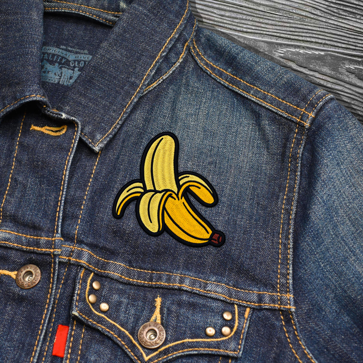 A close-up of a blue denim jacket featuring a yellow embroidered banana patch on the chest, with visible metal buttons and contrast stitching.