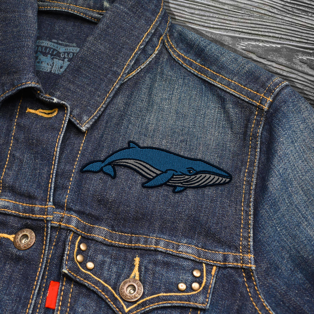 Close-up of a blue denim jacket with a blue whale embroidered patch on the chest, showing yellow contrast stitching, metal buttons, and a gray wood-textured background.