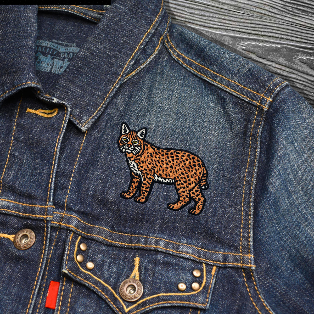Close-up of a blue denim jacket with a colorful embroidered bobcat patch on the chest, featuring yellow contrast stitching and metal buttons.