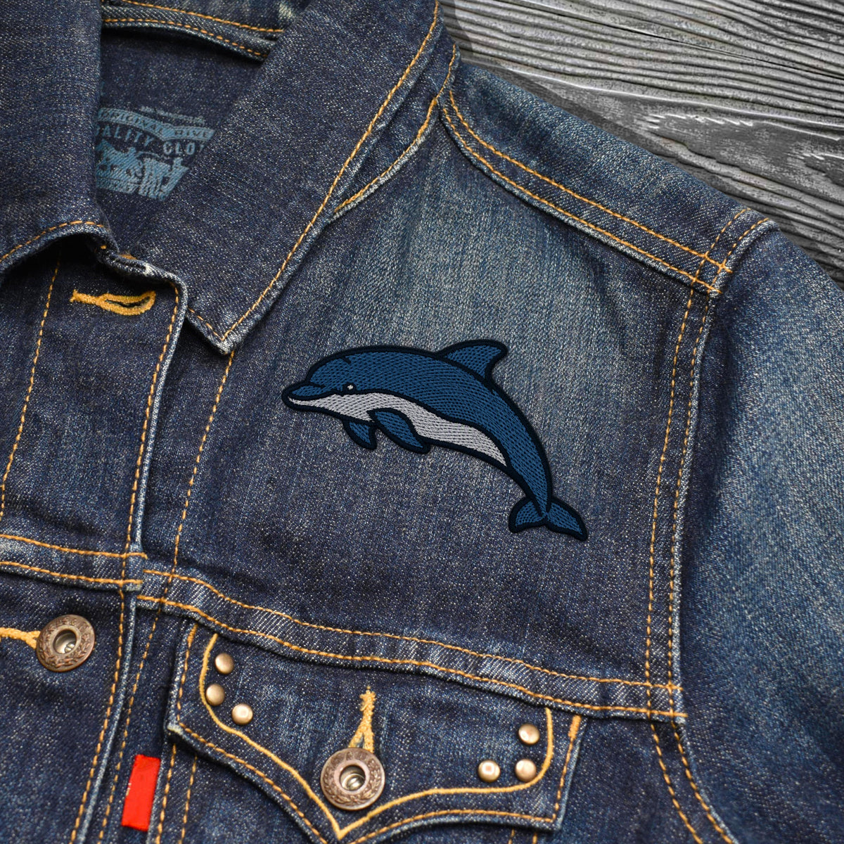 Close-up of a blue denim jacket with a navy and gray embroidered dolphin patch on the chest, shown on a gray wood-textured background.
