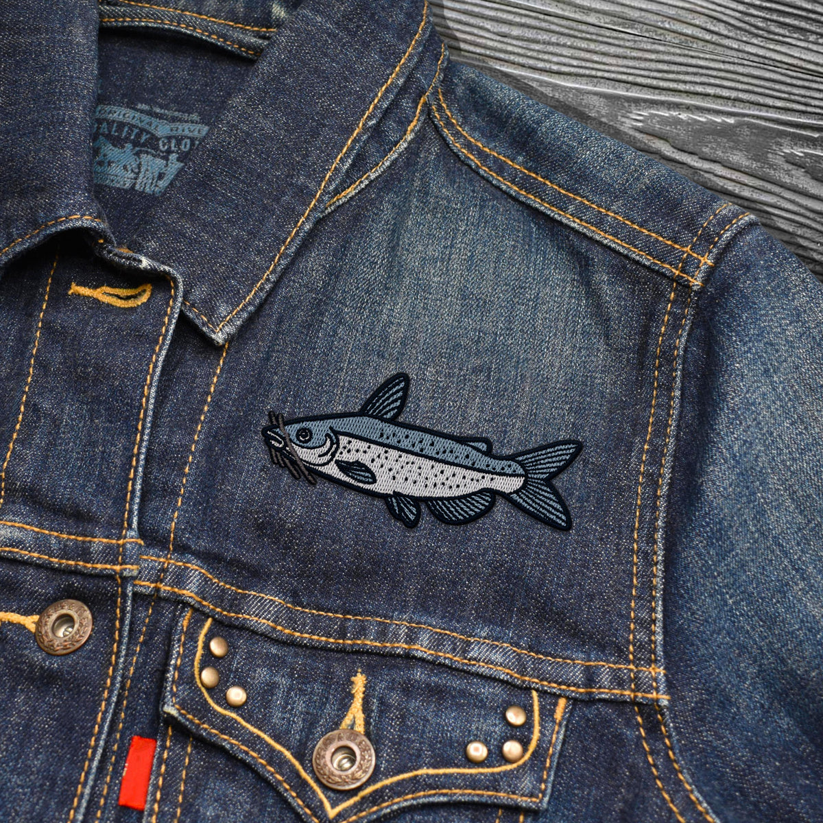Close-up of a dark blue denim jacket with a detailed embroidered channel catfish patch on the chest pocket, featuring yellow contrast stitching and metal snap buttons.