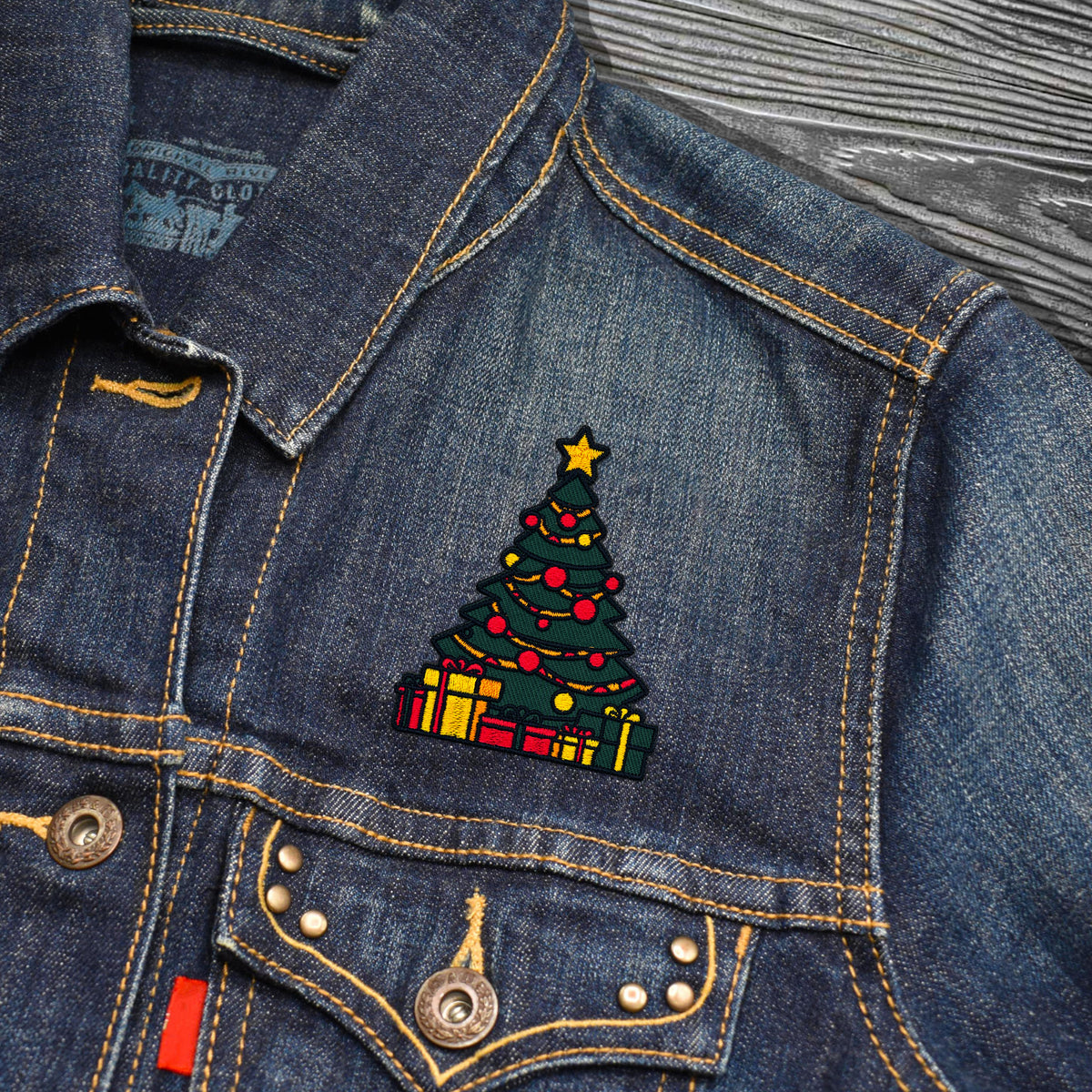 A close-up of a blue denim jacket featuring a colorful embroidered Christmas tree patch with presents on the chest, set against a gray wood-grain background.