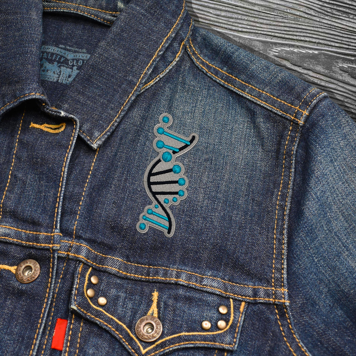 Close-up of a blue denim jacket with a stylized DNA double helix patch in black and turquoise on the chest, showing yellow contrast stitching, metal buttons, and a gray wood-textured background.