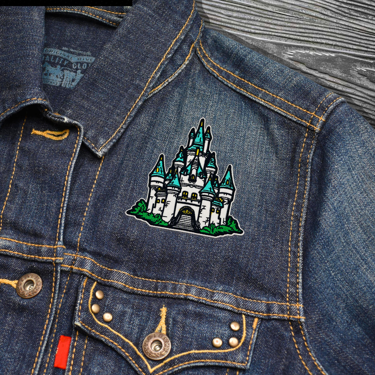 Close-up of a blue denim jacket with a colorful embroidered castle patch on the chest pocket, showing yellow contrast stitching and metal buttons.