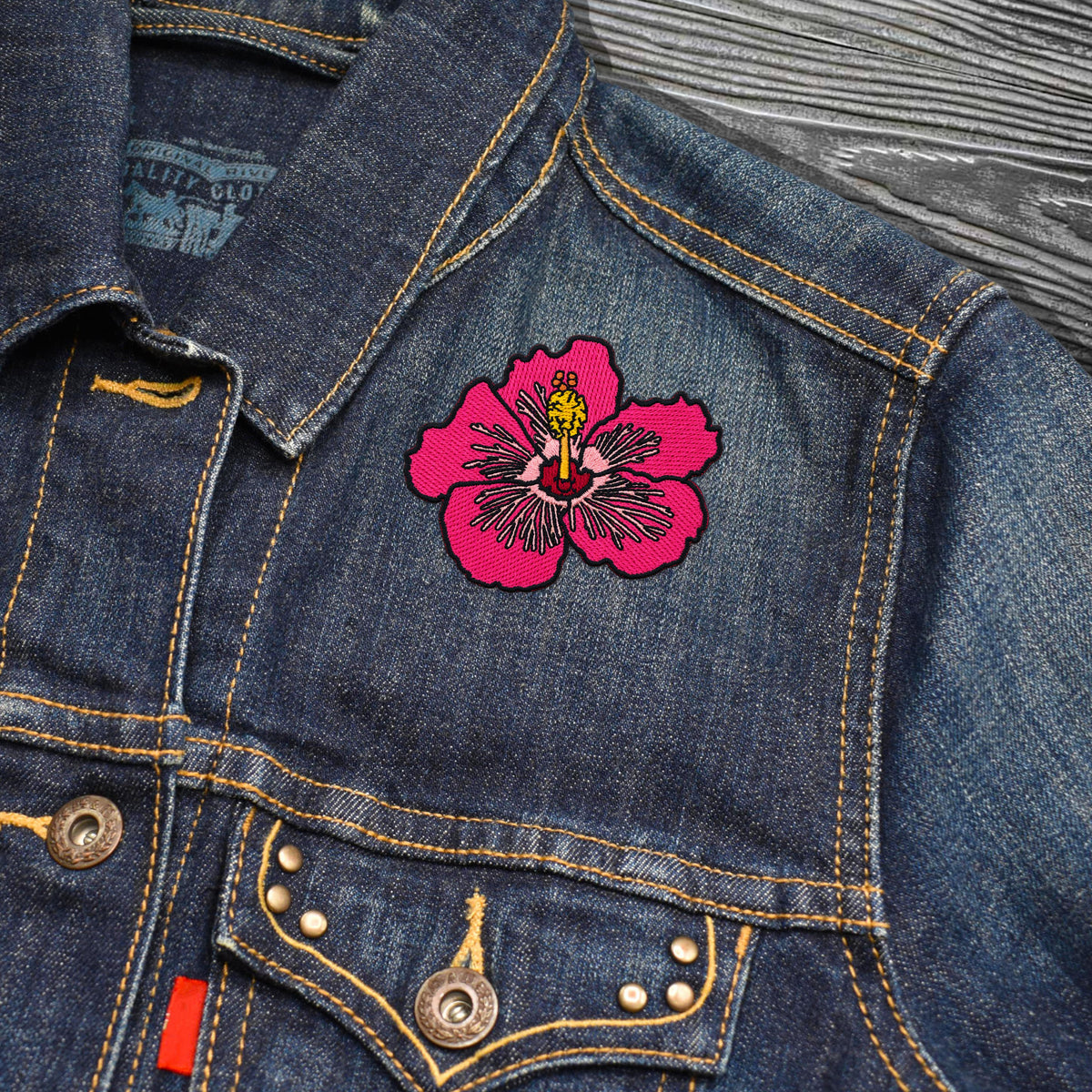 A close-up of a blue denim jacket featuring a vibrant pink embroidered hibiscus flower patch with yellow and red details on the chest, set against a dark gray wood background.