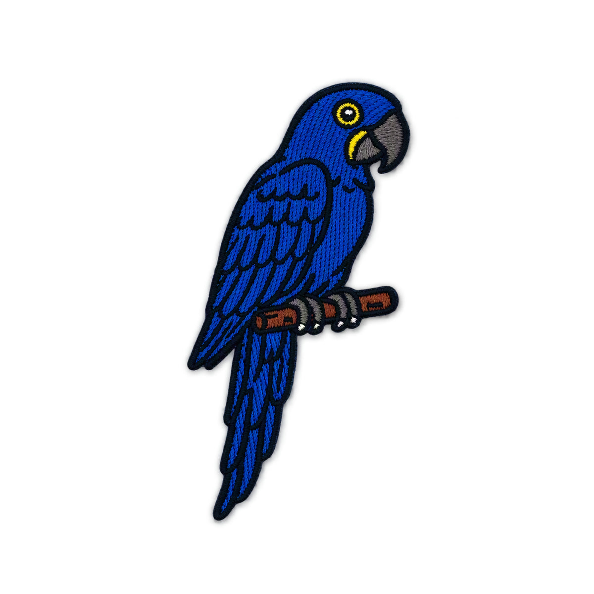 An embroidered patch of a blue parrot perched on a brown branch, featuring a yellow eye and gray beak, isolated on a white background.
