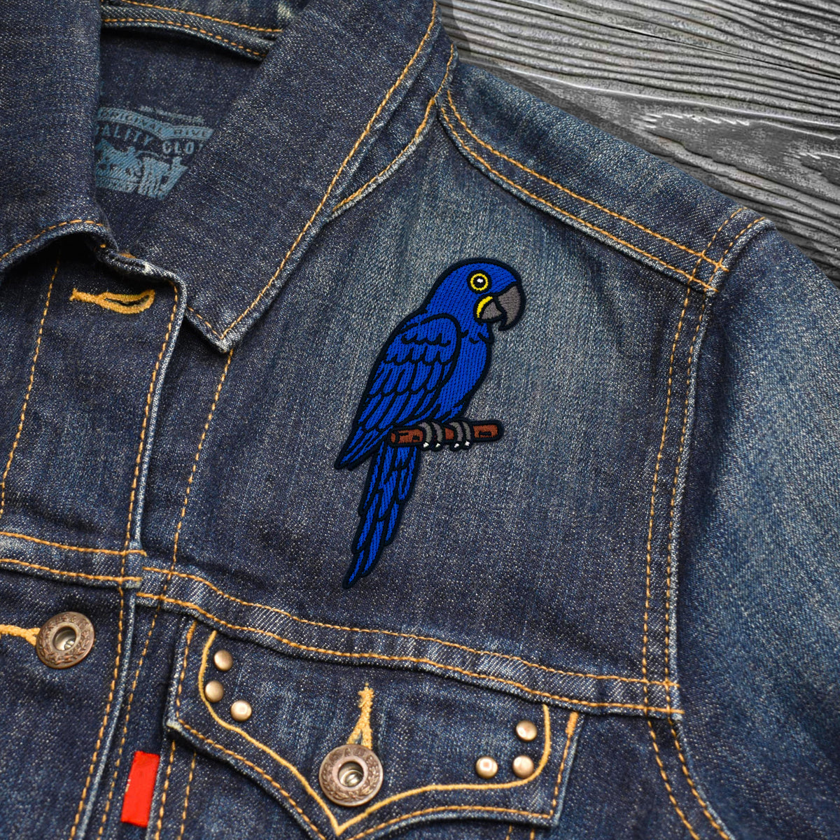 A close-up of a blue denim jacket featuring a vibrant embroidered blue parrot patch on the chest, resting on a gray wood-textured background.