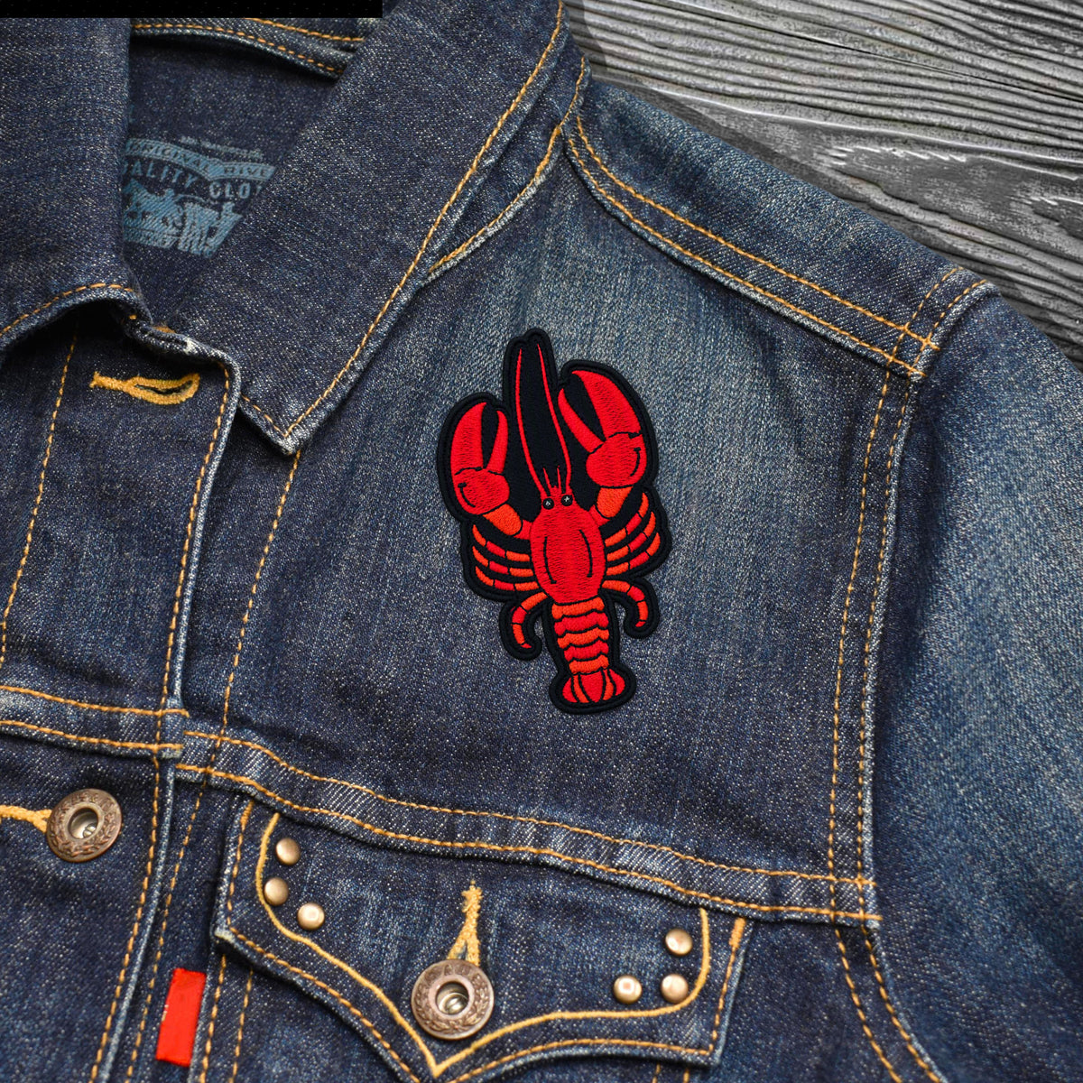 A close-up of a blue denim jacket featuring a red embroidered lobster patch on the chest pocket. The jacket has yellow contrast stitching, metal buttons, and a textured gray wood background.