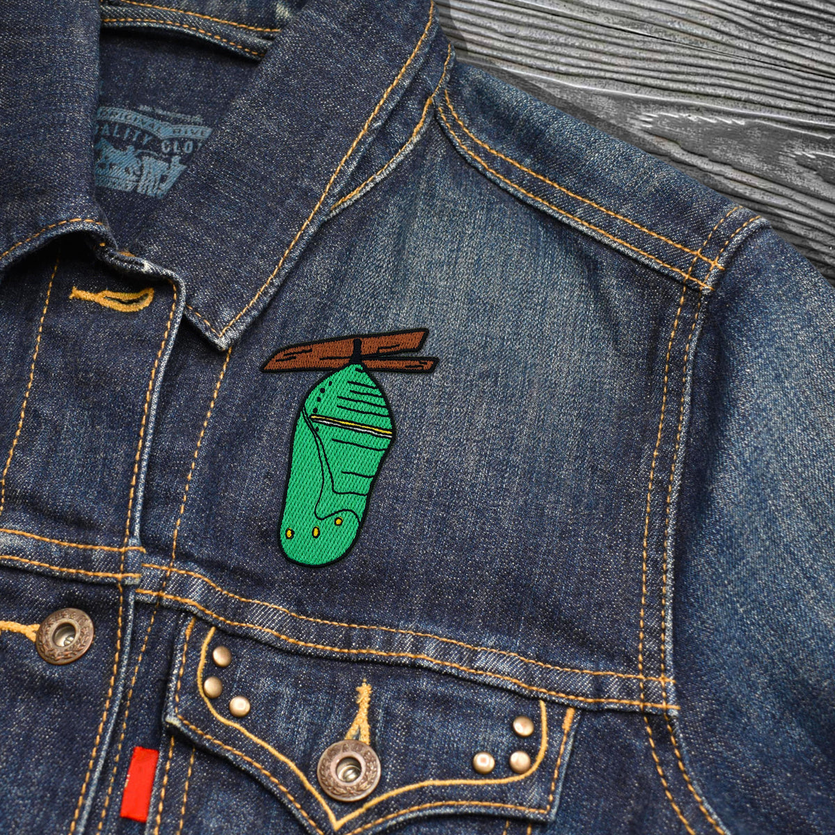 A close-up of a blue denim jacket featuring a green embroidered patch shaped like a boot with a brown wooden top, displayed on a textured gray wood background.