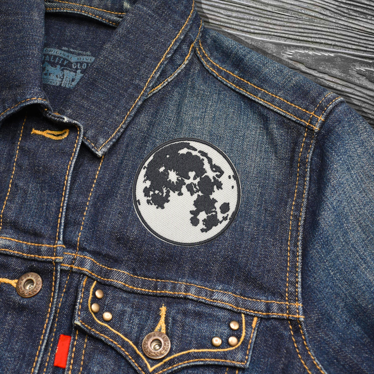 Close-up of a blue denim jacket with a circular moon patch on the chest, showing yellow contrast stitching, metal buttons, and a textured gray wood background.