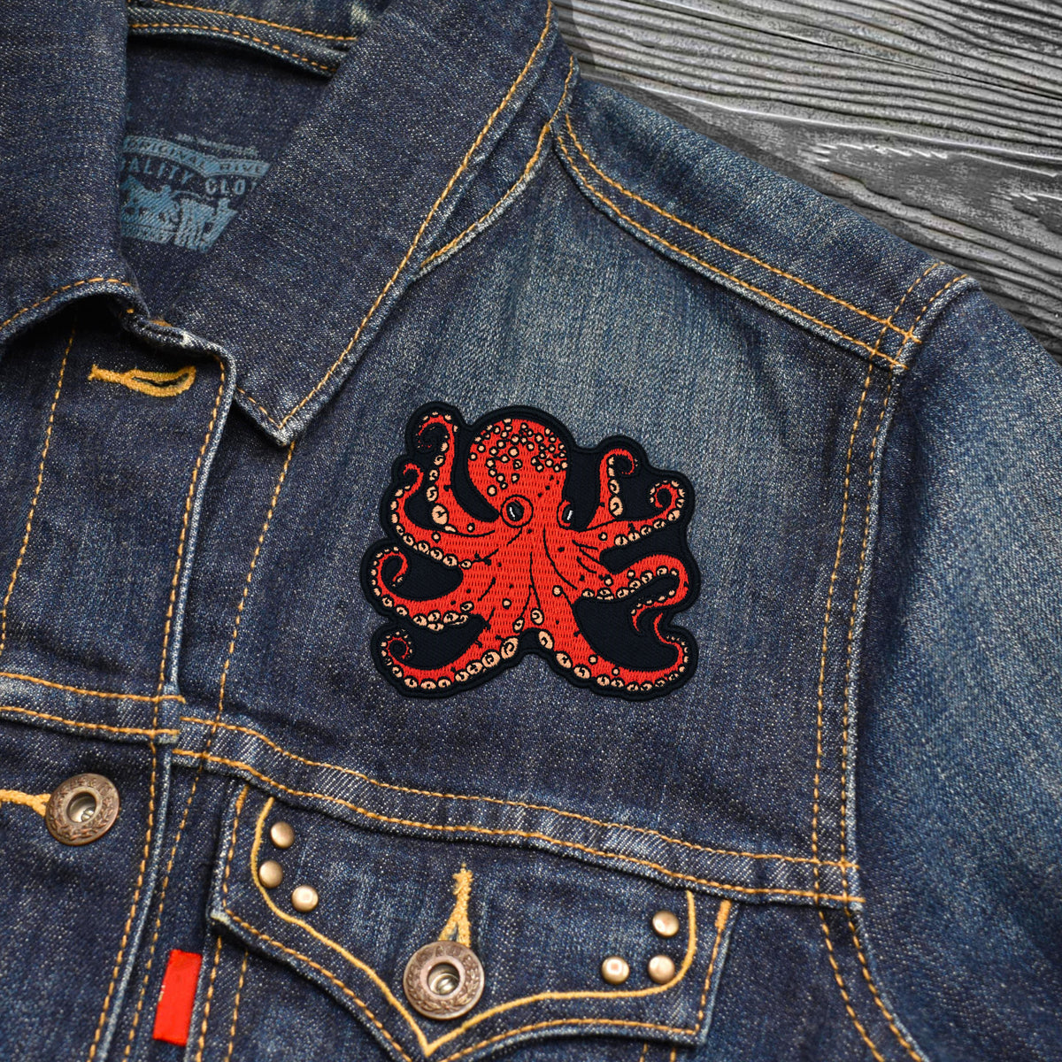 A close-up of a blue denim jacket featuring a prominent red and black embroidered octopus patch on the chest pocket, with gold-toned contrast stitching and metal buttons.