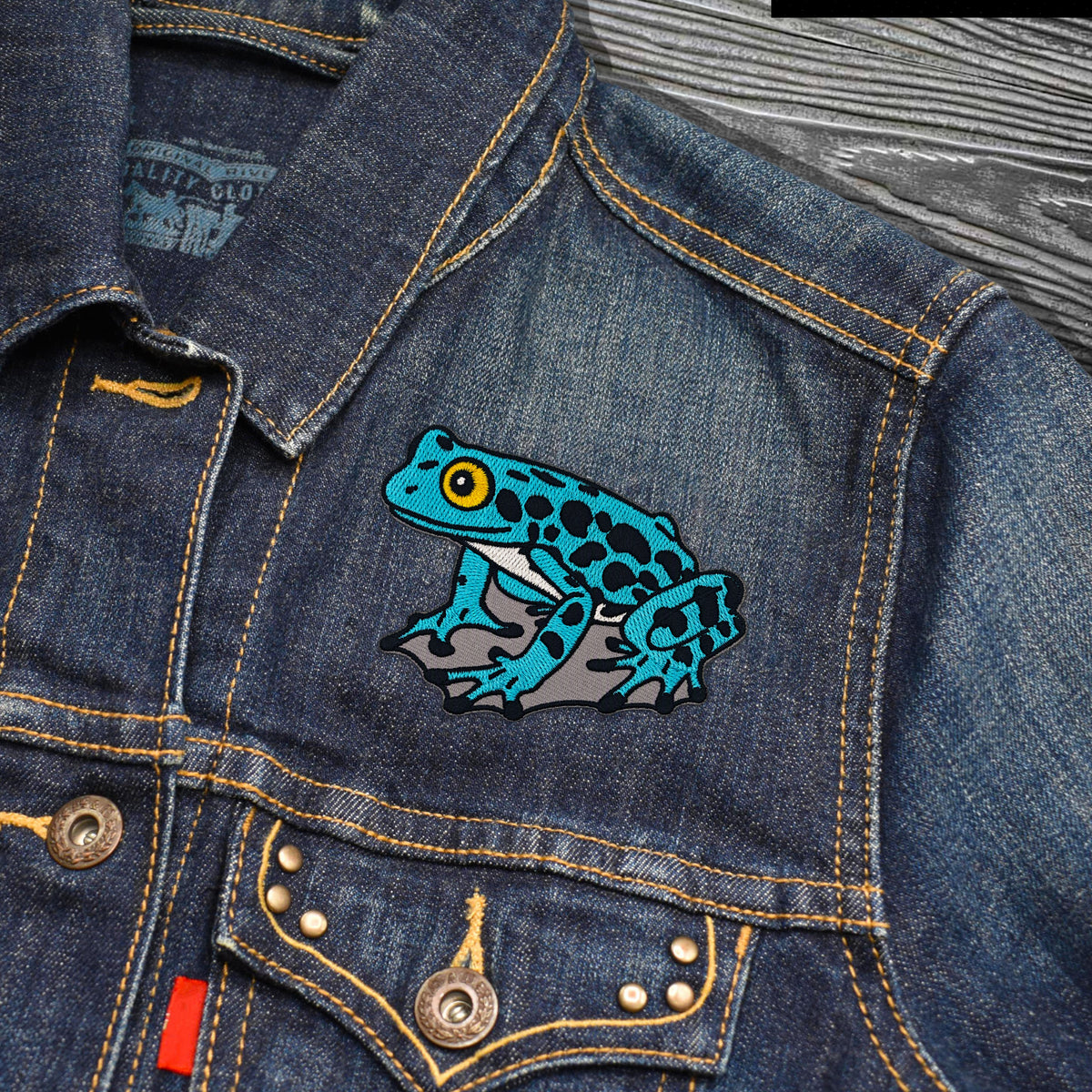 A close-up of a blue denim jacket featuring a colorful embroidered frog patch on the chest pocket. The patch depicts a bright turquoise frog with black spots, yellow eyes, and gray limbs.