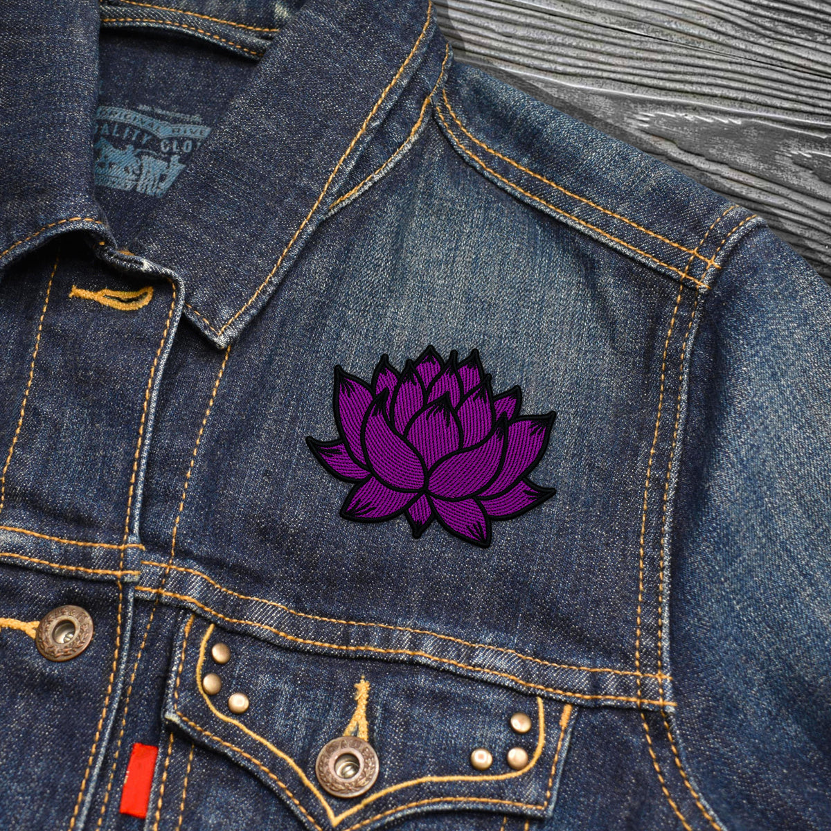 A close-up of a blue denim jacket featuring a purple embroidered lotus flower patch on the chest pocket, with brass buttons and contrast yellow stitching.