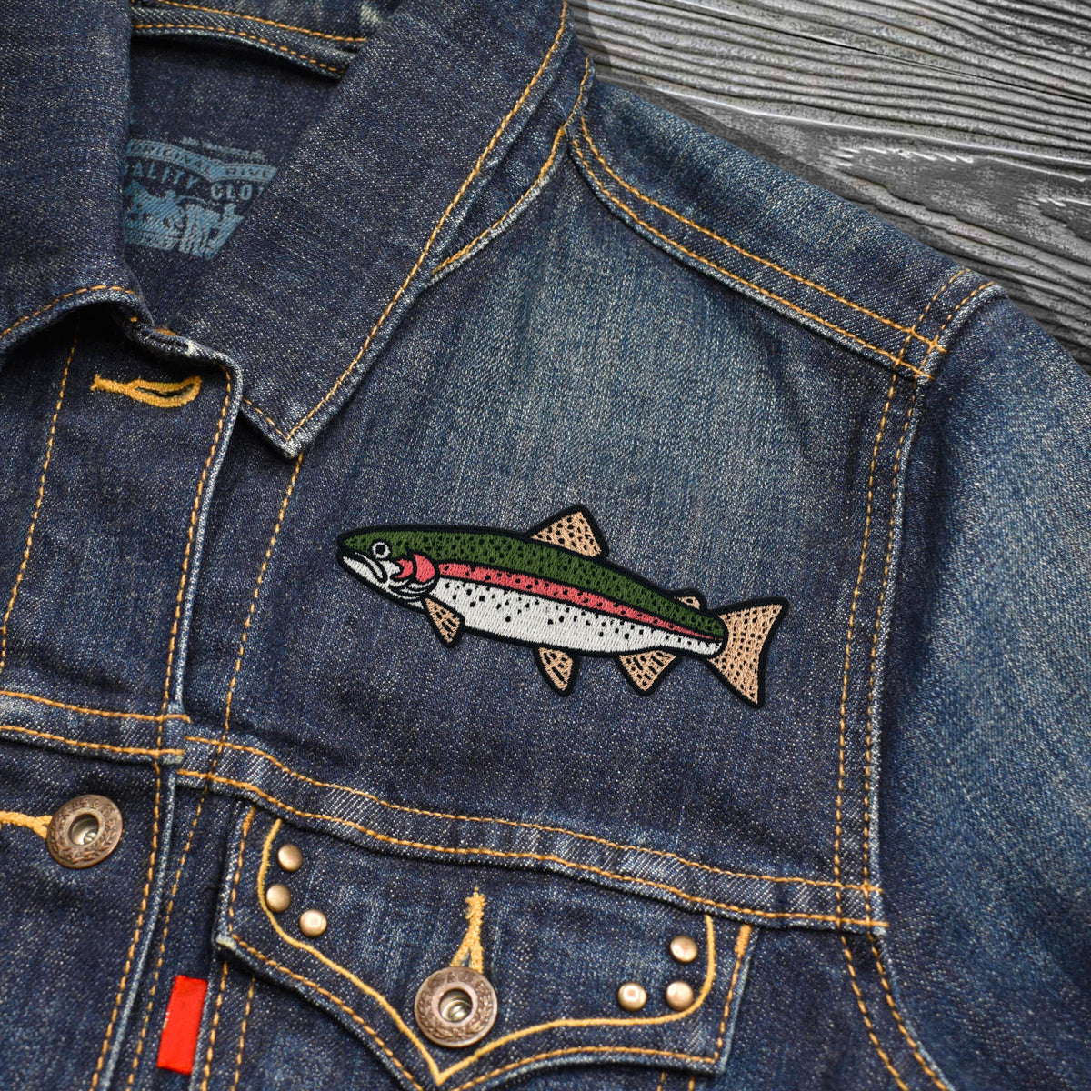 A close-up of a blue denim jacket featuring an embroidered rainbow trout patch on the chest pocket, with visible brass buttons and contrast yellow stitching.