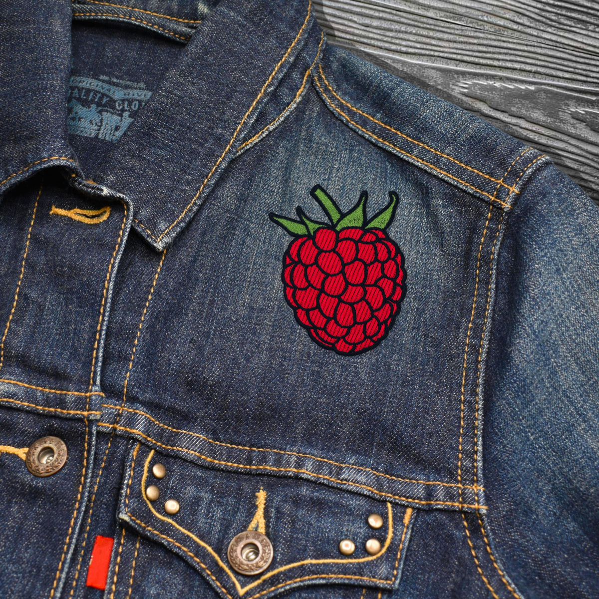 Close-up of a blue denim jacket with a colorful embroidered raspberry patch on the chest, featuring yellow contrast stitching and metal buttons.