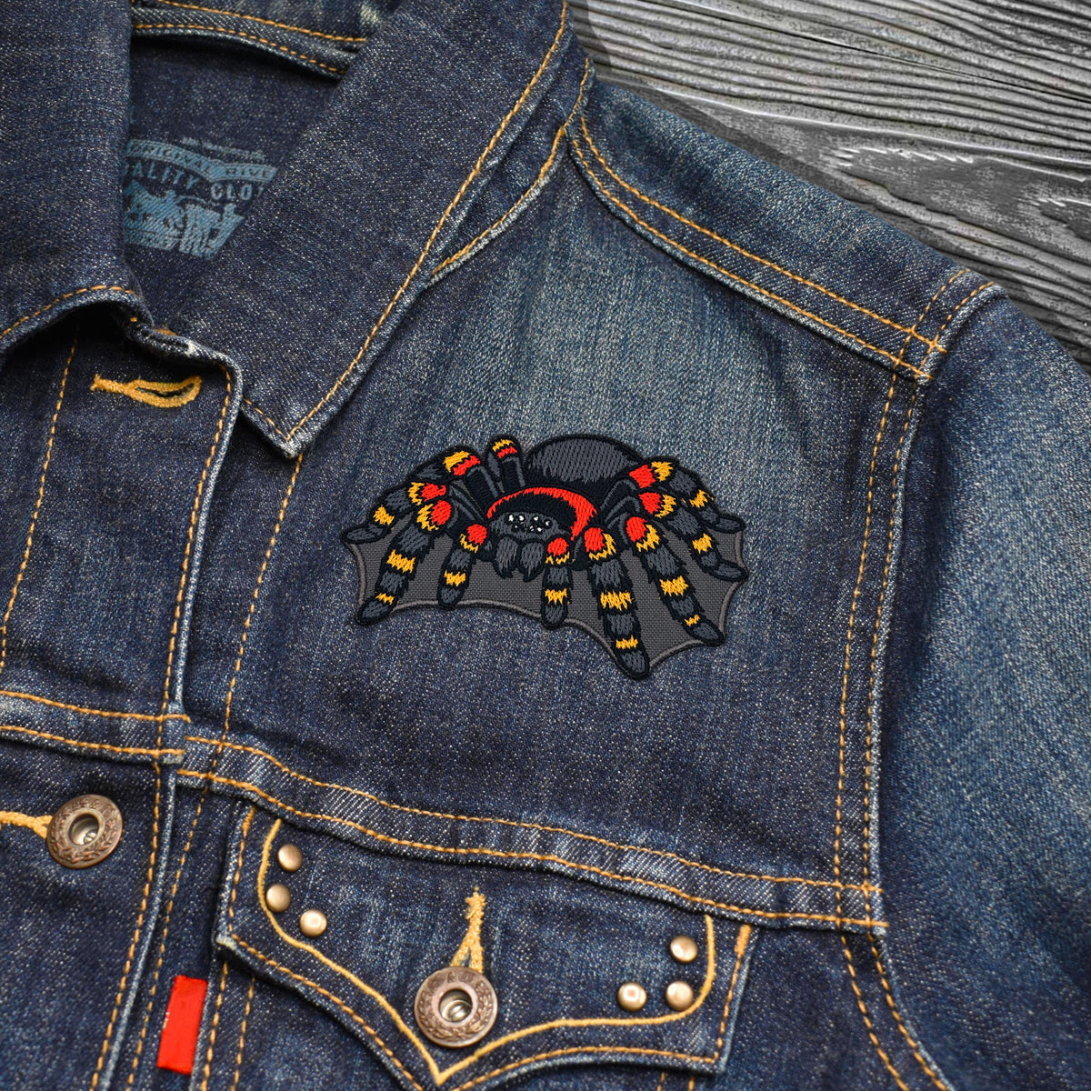 A close-up of a dark blue denim jacket featuring a colorful embroidered spider patch on the chest pocket. The jacket has yellow contrast stitching, metal snap buttons, and a gray wood-textured background.