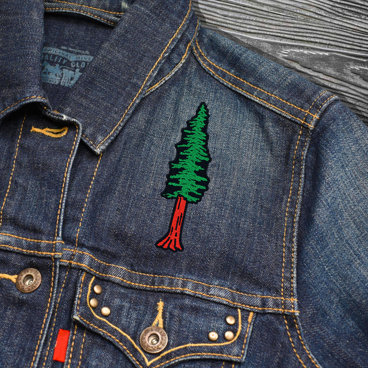 Close-up of a blue denim jacket with a green and red embroidered pine tree patch on the chest, shown on a textured gray wood background.