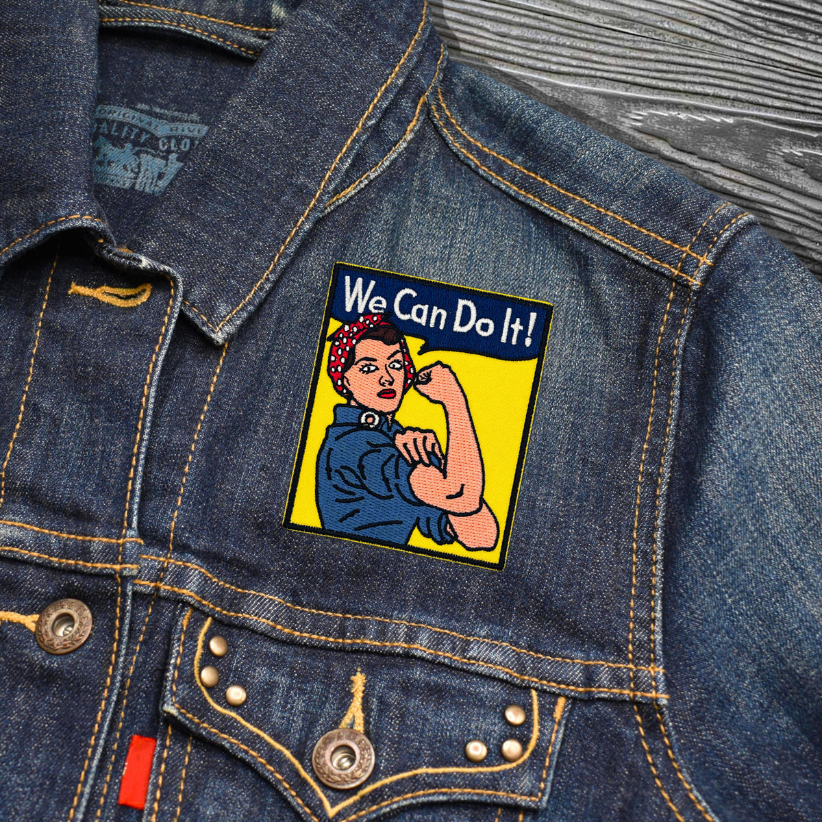 A close-up of a blue denim jacket featuring a rectangular embroidered patch on the chest. The patch depicts the classic Rosie the Riveter illustration with the text 'We Can Do It!' in white on a dark blue banner, set against a bright yellow background.