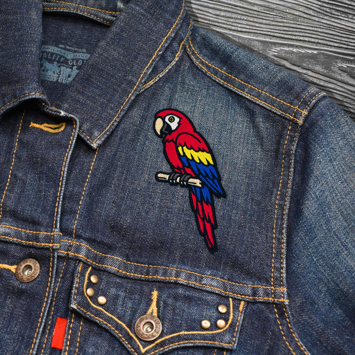A close-up of a blue denim jacket featuring a colorful embroidered parrot patch on the chest, with visible buttons, pockets, and contrast stitching on a textured gray wood background.