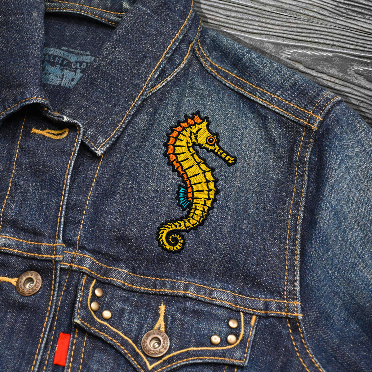 A close-up of a blue denim jacket featuring a colorful embroidered yellow seahorse patch with orange, red, and blue details on the chest, set against a dark wood background.