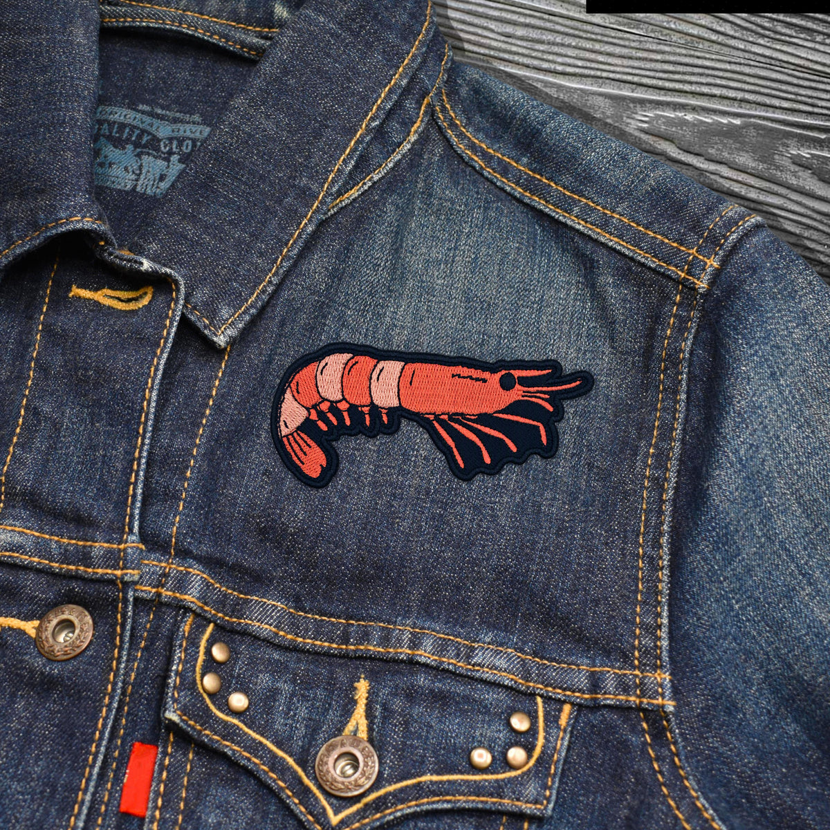 A close-up of a blue denim jacket featuring a prominent embroidered red and orange prawn patch on the chest, with yellow contrast stitching and metal buttons.