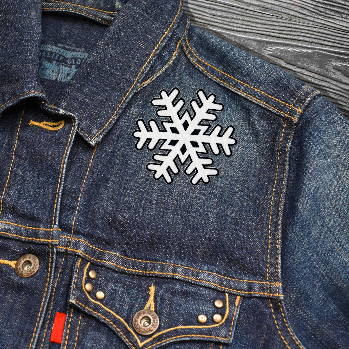 A close-up of a dark blue denim jacket featuring a white snowflake applique with a black outline on the chest, set against a gray wood-grain background.
