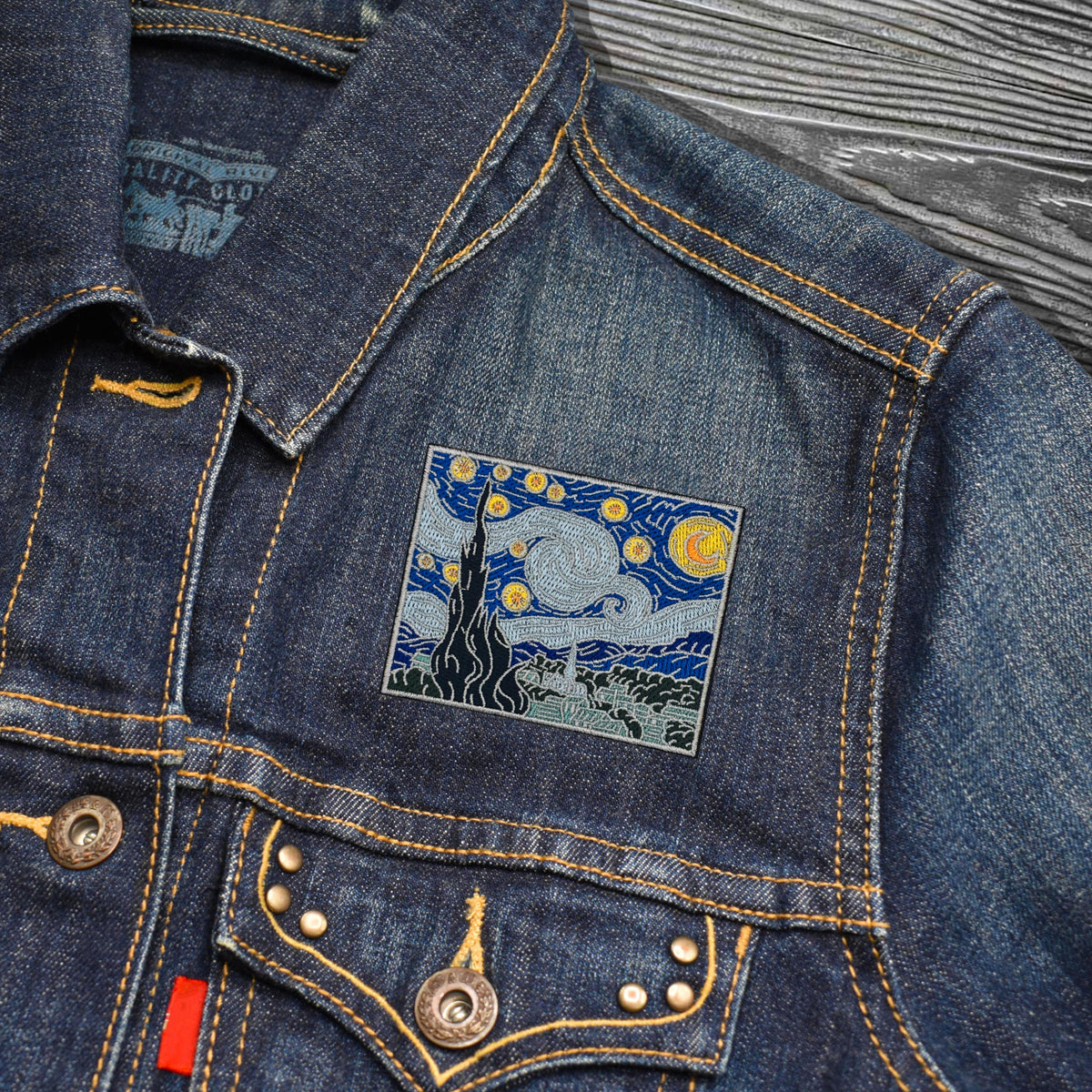 A close-up of a blue denim jacket featuring a square embroidered patch on the chest with a Van Gogh-inspired night scene in blue, yellow, and black. The jacket has yellow contrast stitching, metal buttons, and a red tab on the pocket, photographed against a gray wood-textured background.