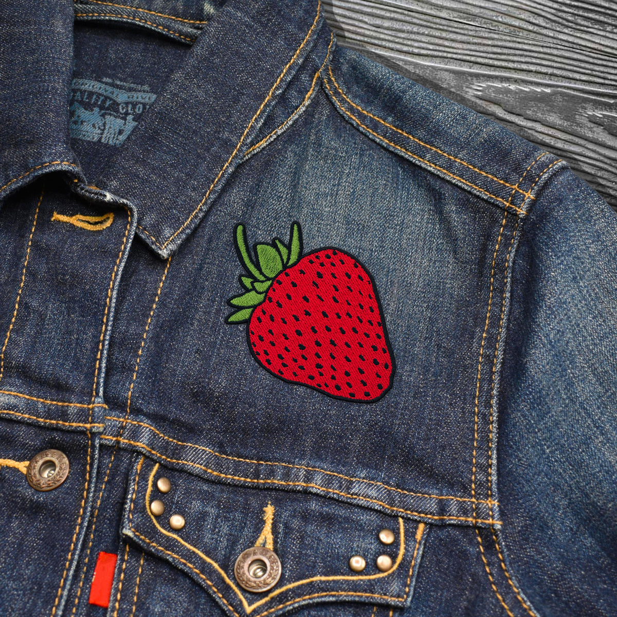 Close-up of a blue denim jacket with a large embroidered red strawberry patch on the chest, featuring yellow contrast stitching and metal buttons.
