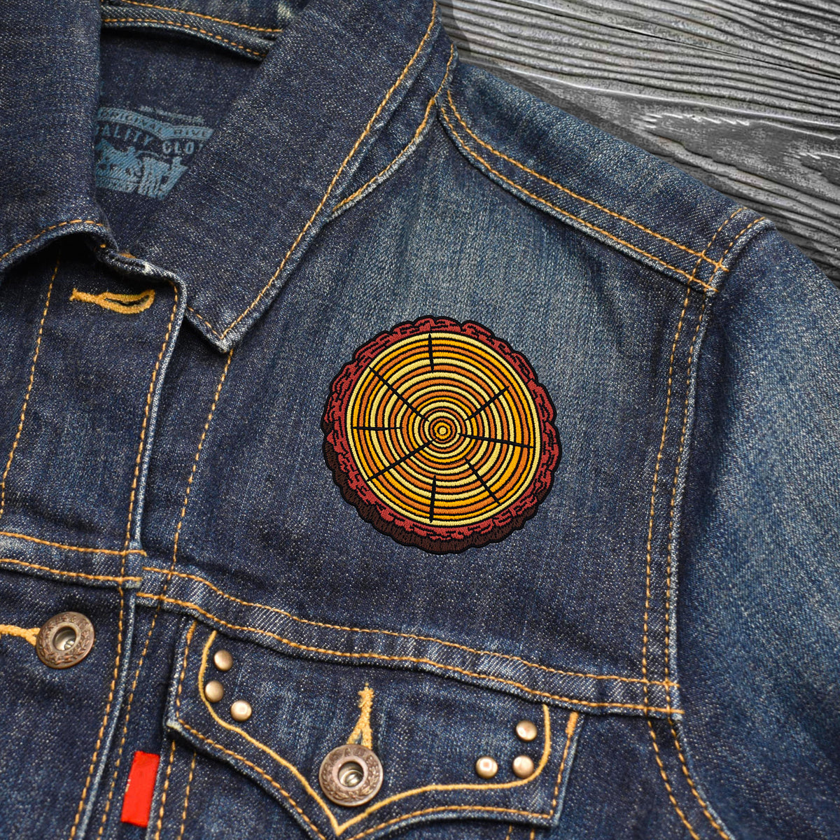 A close-up of a blue denim jacket featuring a circular embroidered patch with concentric yellow, orange, and red rings on the chest, set against a gray wood-textured background.