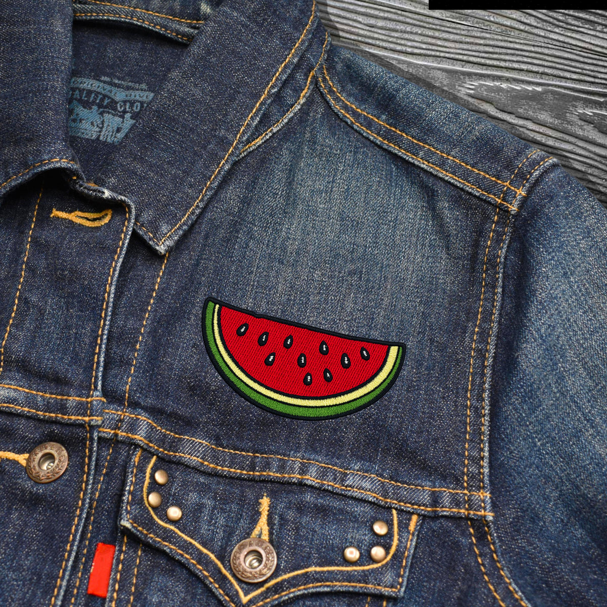 Close-up of a blue denim jacket with a colorful embroidered watermelon patch on the chest pocket, showing yellow stitching and metal buttons.